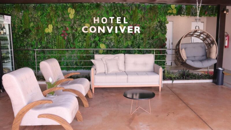 Recepcão do hotel, onde se vê um sofá, duas poltronas, uma poltrona suspensa e ao fundo um painel com folhas escrito hotel coniver